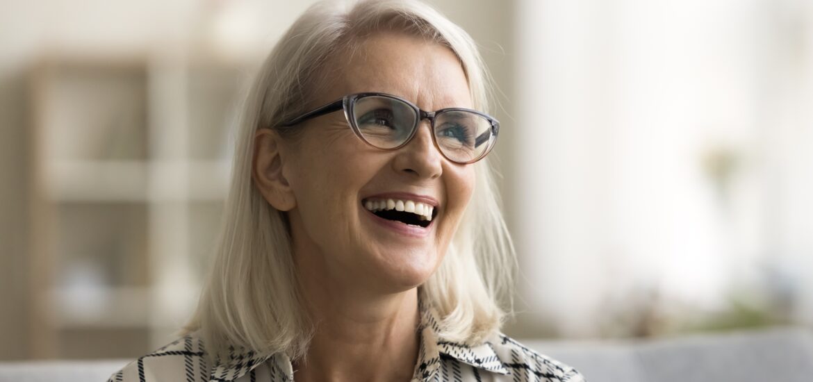 Happy beautiful blonde mature woman in elegant glasses sitting on couch, looking away, smiling, laughing with open mouth, showing healthy white teeth, dental implants